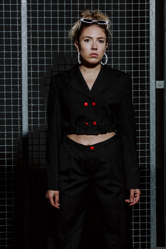 Fashion, Frau in schwarzem bauchfreien Nadelstreifenanzug mit roten Knöpfen und weißer Sonnenbrille vor Metallgitter, Halbaufnahme, Hochformat