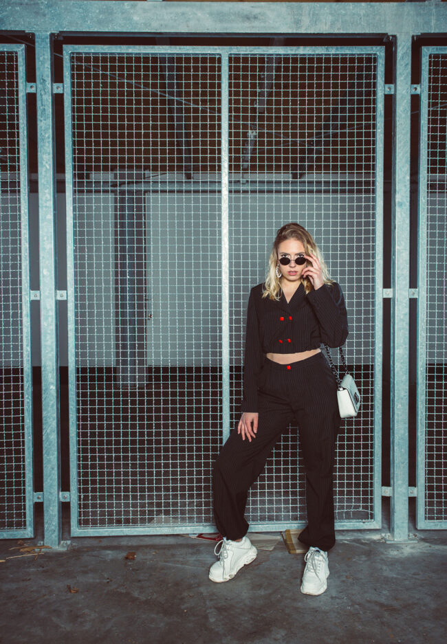 Schwarz gekleidetes Model mit weißen Schuhen steht vor einem Metallgitter Tor