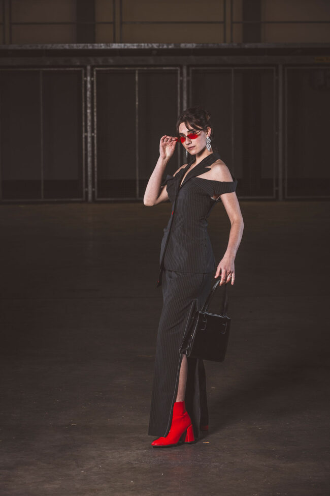 Fashion, Model in schwarzem Kostüm und schwarzer Tasche, mit roten Schuhen und roter Brille in einer dunklen Halle