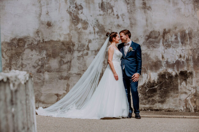Hochzeit, Portarit, Paar hält sich im Arm und schauen sich an, sie stehen vor einer weiß-grauen Betonwand