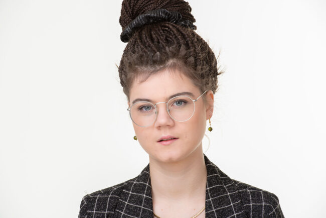 Portrait, Frau mit Dead-Locs, Brille und Blazer schaut ernst in die Kamera