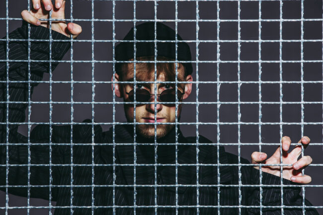 Portrait Junger Mann mit Sonnenbrille und dunkler Kleidung hinter einem Metallgitter