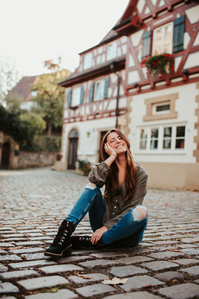 Portrait, Frau sitzt auf Kopfsteinpflaster vor Fachwerkhaus und strahlt in die Kamera, Ganzaufnahme