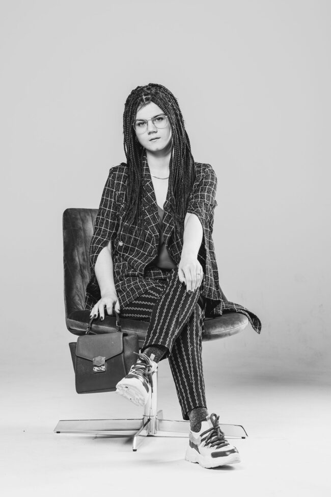 Portrait, Frau mit Blazer und Tasche in der Hand sitzt auf Sesselkante, heller Hintergrund, Ganzaufnahme, Schwarz-Weiß