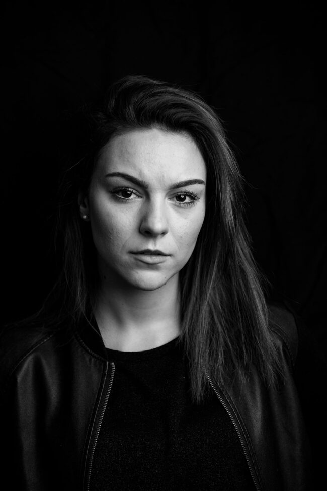 Portrait, Frau in dunkler Jacke schaut ernst in die Kamera, schwarzer Hintergrund