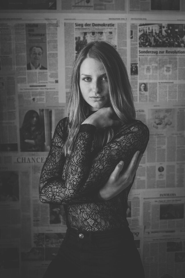 Portraitshooting, junge Frau in Spitzenbluse vor Zeitungswand, ernster Blick in die Kamera, Arme vor dem Körper verschränkt, Schwarz-Weiß