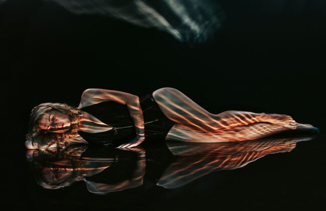 Sensual, Frau in schwarzem Body liegt auf nassem Boden, Wasserspiegelungen reflektieren sich auf dem Körper