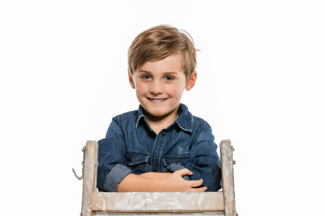 Portrait, kleiner Junge in Jeanshemd lehnt auf Klappleiter und lächelt in die Kamera, Nahaufnahme