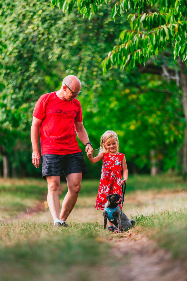 Familie, Vater in rotem Shirt und kleine Tochter in rotem Kleid auf Feldweg zwischen Bäumen, mit Mops-Hund