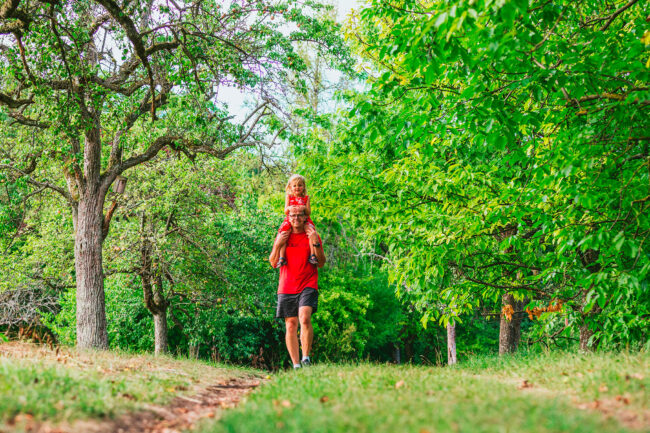 Familie, Outdoor, Vater in rotem Shirt hat Tochter in rotem Kleid auf den Schultern und läuft zur Kamera