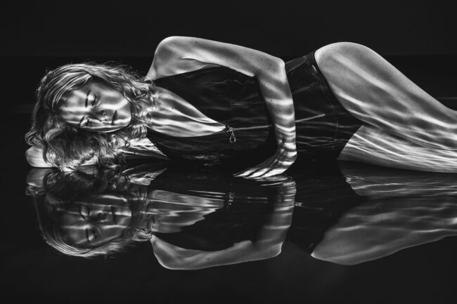 Sensual, Frau in schwarzem Body liegt auf nassem Boden, Wasserspiegelungen reflektieren sich auf dem Körper, Halbportrait, Schwarz-Weiß