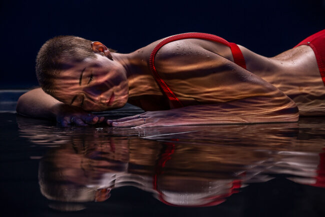 Sensual, Frau in rotem Bikini liegt auf nassem Boden, Wasserspiegelungen reflektieren sich auf dem Körper, Augen geschlossen, Halbportrait