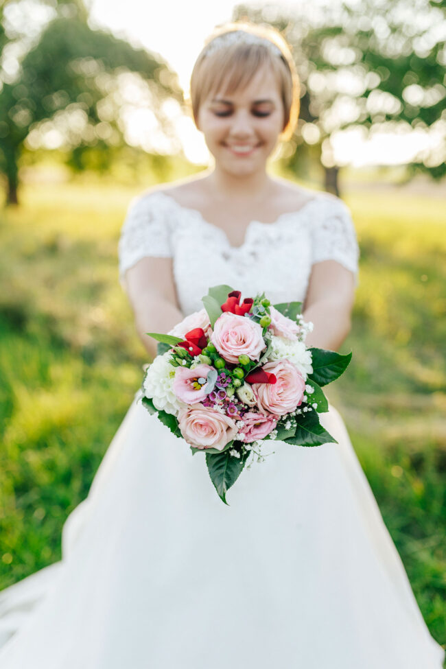 Hochzeit, Portrait, Braut hält Brautstrauß in die Kamera und lacht, in der Dämmerung