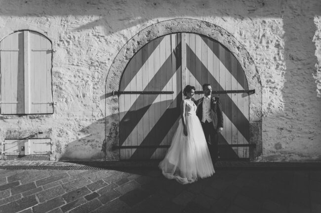 Hochzeit, Portrait, Brautpaar steht in der Abendsonne vor einem doppelflügligen, gestreiften Holztor und schaut in die Ferne, Querformat, schwarz-weiß