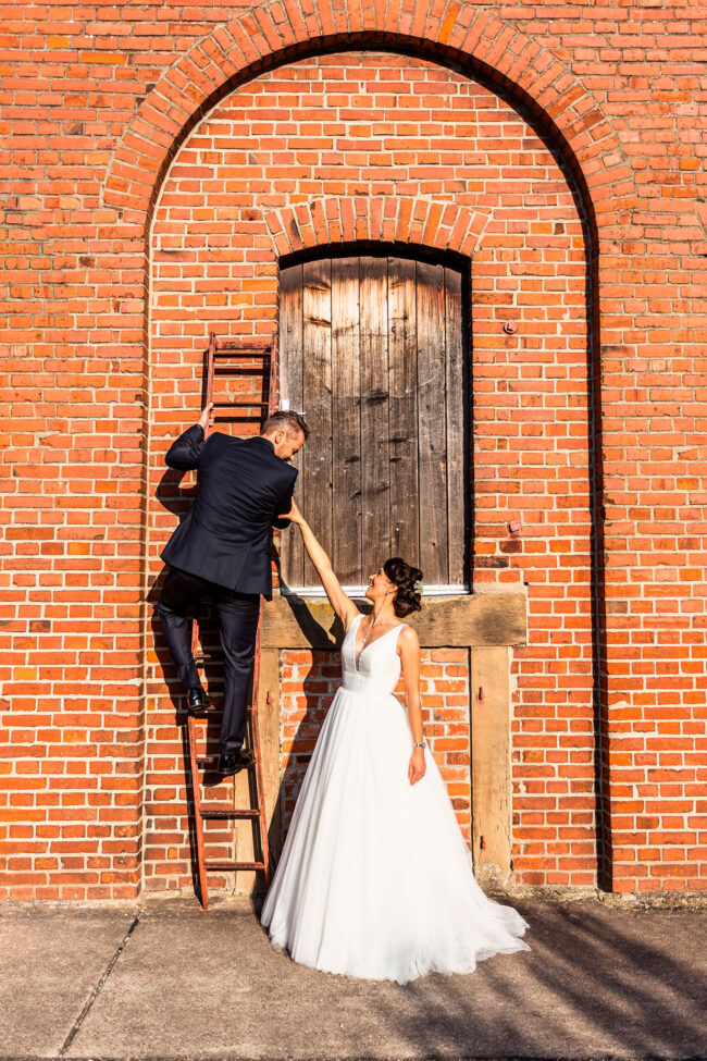 Hochzeit, Bräutigam klettert eine rote Leiter an einer roten Backsteinmauer hoch. Braut hält Mann am Arm fest, Paar schaut sich an