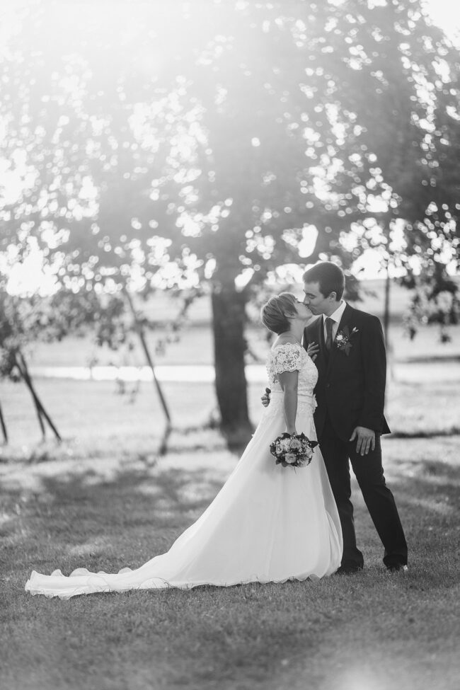 Hochzeit, Ganzaufnahme, Paar steht auf Wiese und küsst sich, durch Baum im Hintergrund scheint die Sonne, schwarz-weiß, Hochformat
