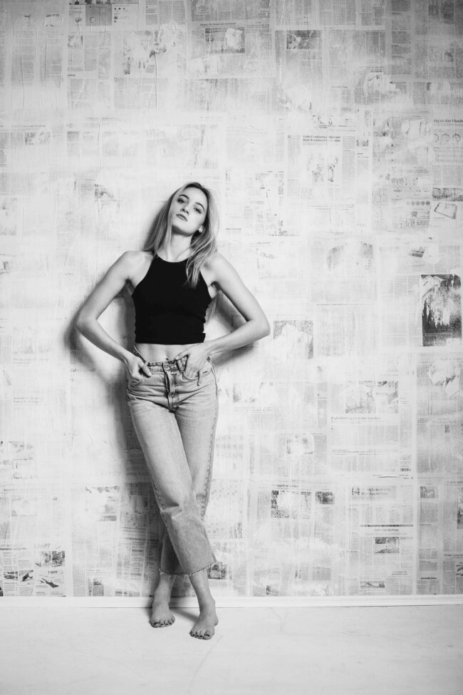 Portrait, junge Frau in schwarzem Oberteil und Jeans mit überkreuzten Beinen vor Zeitungswand stehend, Ganzaufnahme, Schwarz-Weiß