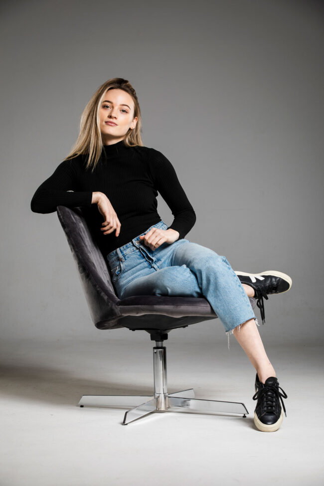 Portrait, junge Frau sitzt mit verschränktem Bein auf grauem Sessel, Ganzaufnahme
