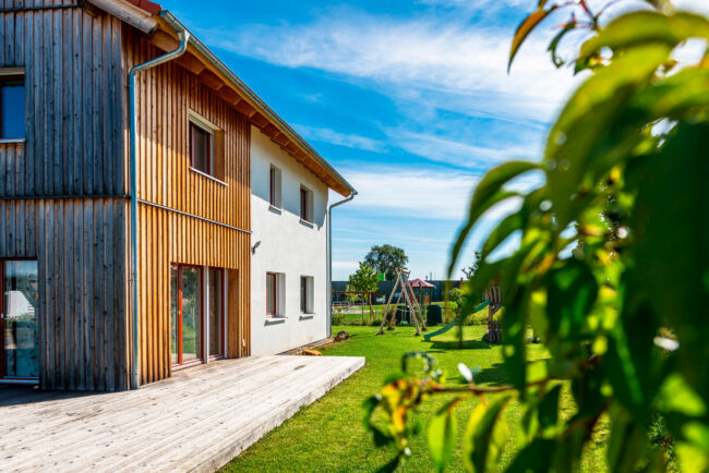 Architektur, Teilaufnahme der Hausseite mit Garten und Rasenfläche. Haus ist zur Hälfte weiß verputzt und die 2. Hälfte ist mit vertikaler Holzverkleidung