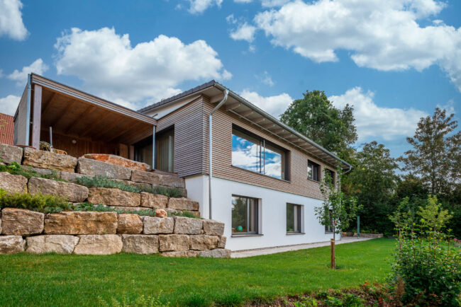 Architektur, Rückansicht weißes Haus, OG mit horizontaler Holzverkleidung, linkt ist eine Natursteinmauer, davor eine Rasenfläche
