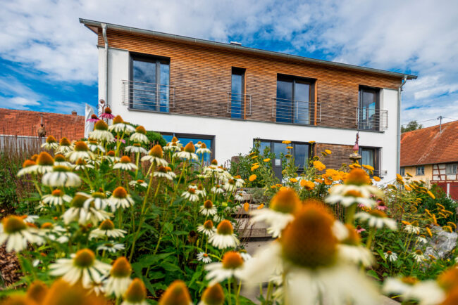 Architektur, Aufnahme der Rückansicht eines weisen Hauses mit Teilholzverkleidung, im Vordergrund stehen Kornblumen im Garten