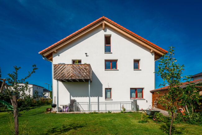 Architektur, weiße Hausseite mit kleinem Holzbalkon. Rote Fensterrahmen, davor eine Grüne Rasenfläche und zwei kleinen Bäumen