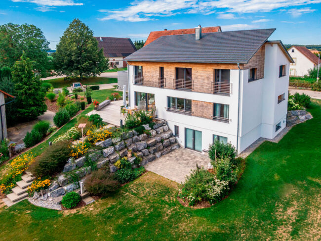 Architektur, modernes dreistöckiges Wohnhaus mit weißer Fassade und Holz, Solarplatten auf dem Dach, mit großem Garten, Drohnenaufnahme