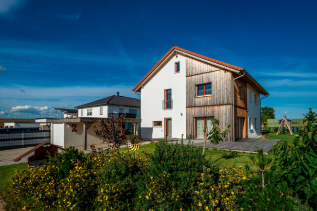 Architektur, Wohnhaus mit weißer Fassade mit Holz, im Vordergrund Grünpflanzen und ein grüner Garten, vor blauem Himmel