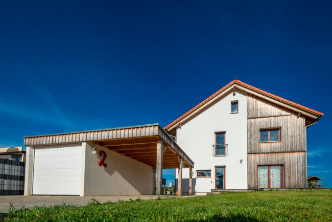 Architektur, Wohnhaus mit weißer Fassade und Holz mit grünem Garten und Garage vor blauem Himmel