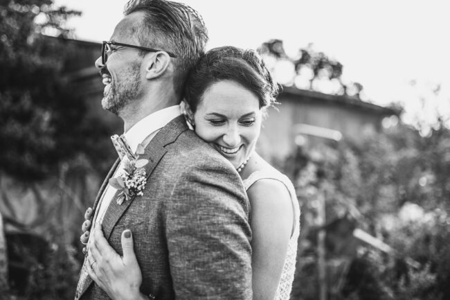 Hochzeit, Portrait, Outdoor, Braut umarmt Bräutigam von hinten und beide lachen, Querformat, schwarz-weiß