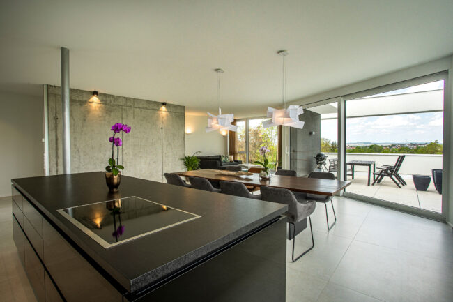 Interieur Küche mit freistehender schwarzer Küchenzeile, im Hintergrund Essbereich mit großem Tisch, dahinter Balkon mit Sitzgelegenheit