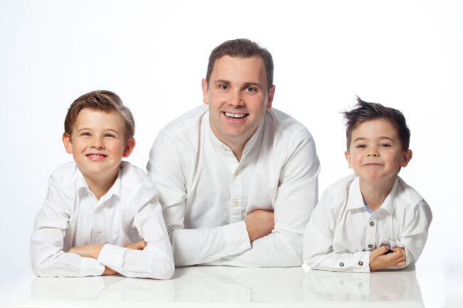 Familienshooting, Vater mit zwei jungen Söhnen in weißen Hemden auf weiße Tischplatte aufgelehnt, weißer Hintergrund, Querformat