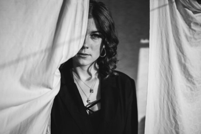 Protrait, Frau mit schwarzem Oberteil schaut neutral in die Kamera, halbes Gesicht von weißem Stoff verdeckt, Halbportrait, Schwarz-Weiß