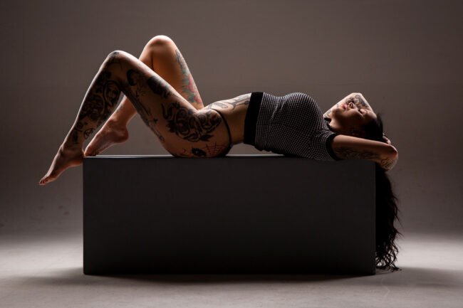 sensual, Frau in schwarz-weißem Shirt und schwarzer Wäsche liegt auf dem Rücken mit angewinkelten Beinen auf Podest, beide Arme über dem Kopf, Augen geschlossen, hat viele Tätowierungen am Körper