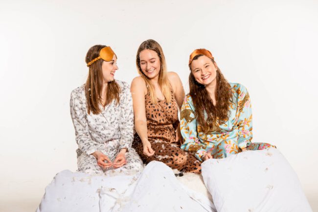 Freundschaftsshooting, drei Frauen in Pyjamas sitzen zwischen Kissen und Federn und lachen in die Kamera, weißer Hintergrund