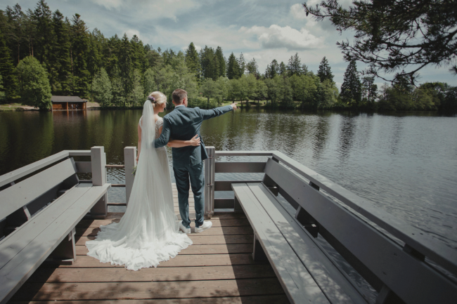 Hochzeit, Portrait, Brautpaar auf Steg, Rückenansicht, Bräutigam zeigt in die Ferne