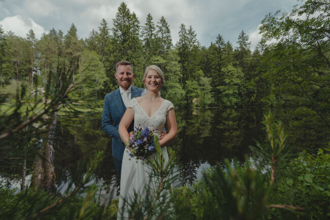 Hochzeit, Brautpaar vor See in grüner Naur, beide lächeln in die Kamera