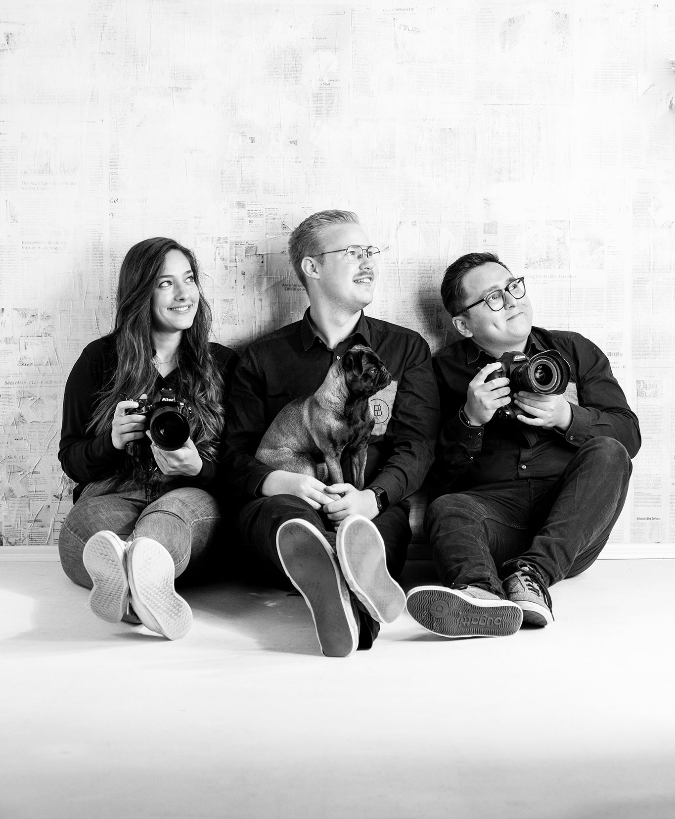 Team von BE fotomedia an Wand sitzend, mit Kameras und Hund, Schwarz-Weiß