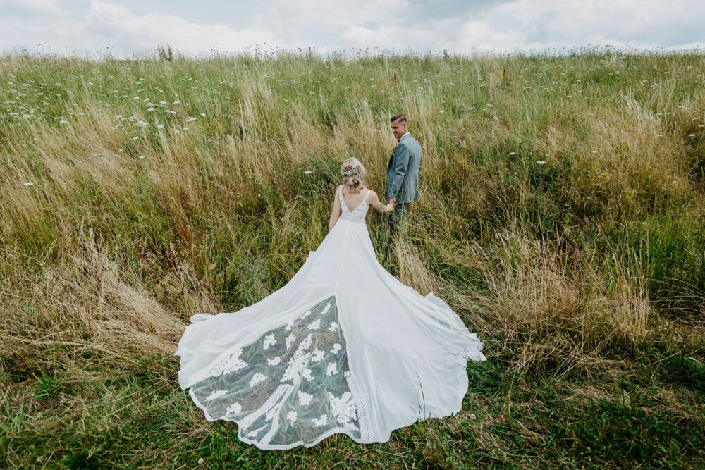 Hochzeit, Brautpaar in grünem Feld, Ganzkörperaufnahme, sich an den Händen haltend, Braut mit Rücken zur Kamera, Querformat