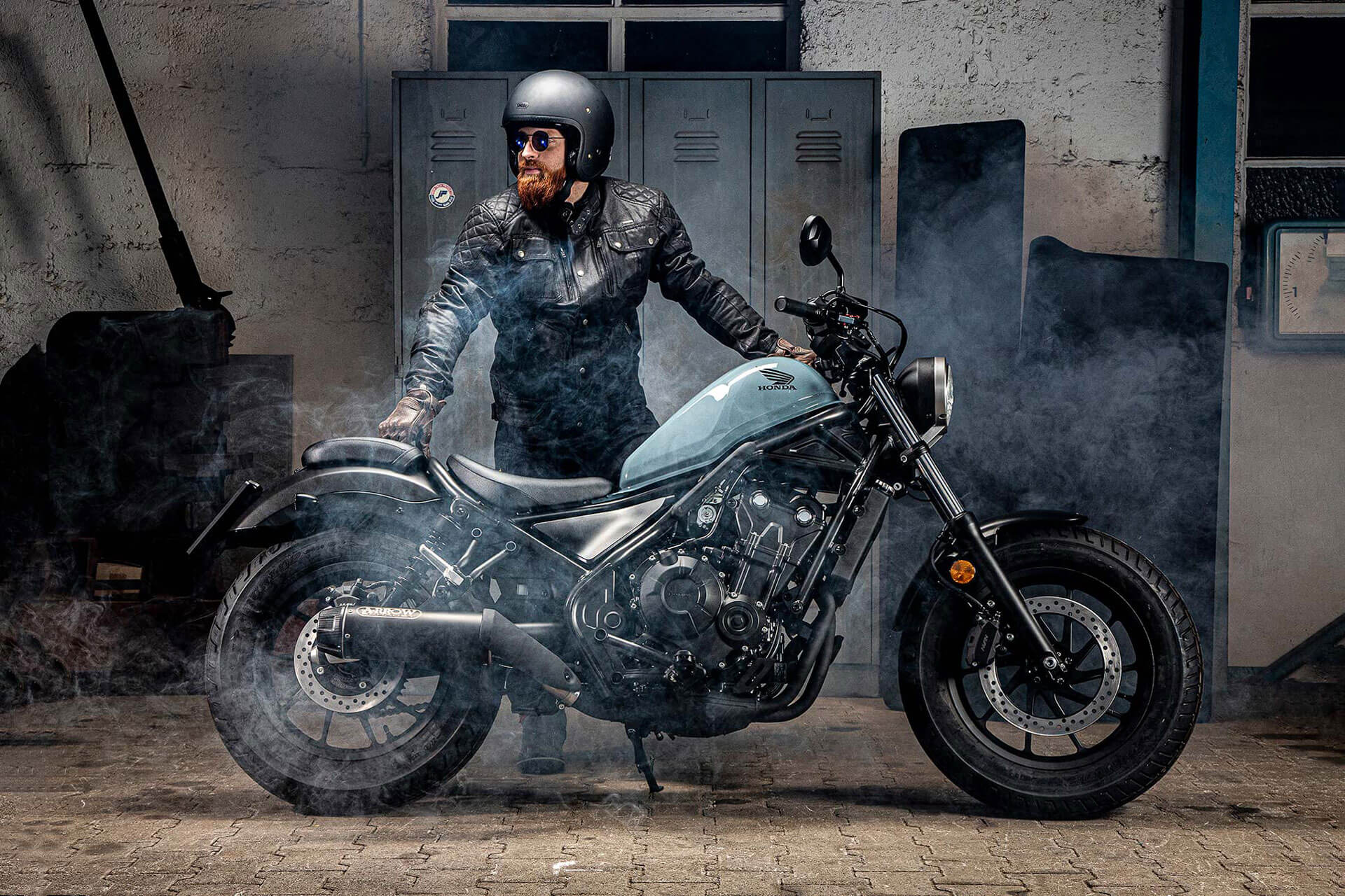 Motorradfahrer steht neben einem schwarzen Motorrad. Dabei kommt etwas Nebel von der Seite.