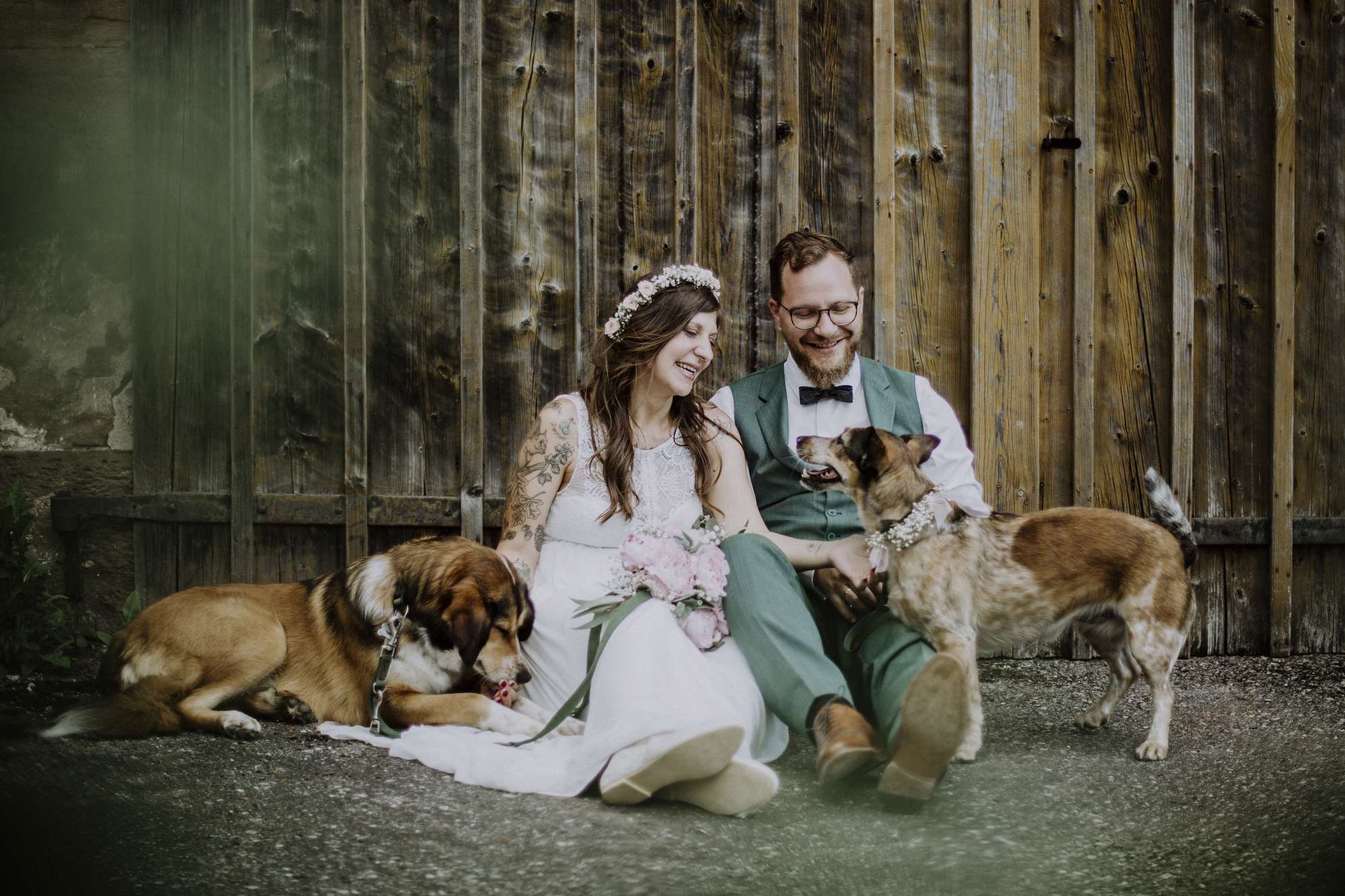 Ein Brautpaar sitzt mit Hunden auf dem Boden vor einer Holzwand.