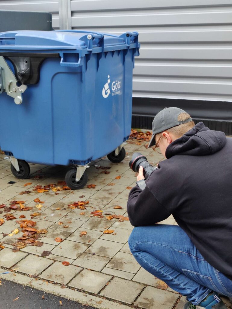Ein Fotograf macht ein Bild von einem blauen Muelleimer auf einer gepflasterten Flaeche. oplus_1048624