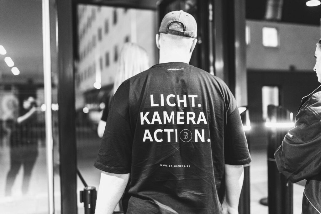 Ein Mann in einem schwarzen Shirt mit der Aufschrift "Licht Kamera Action" geht durch eine Tuer.