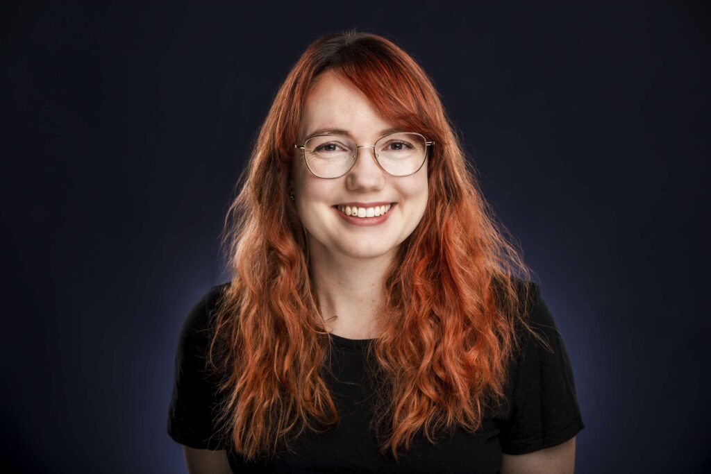 Portraet einer laechelnden Frau mit rotem Haar und Brille im dunklen Hintergrund.