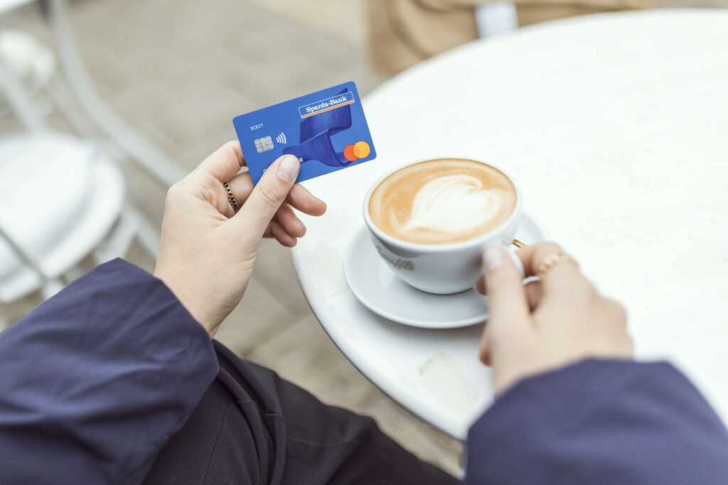 Eine Person haelt eine Kaffeetasse und eine Kreditkarte in den Haenden.