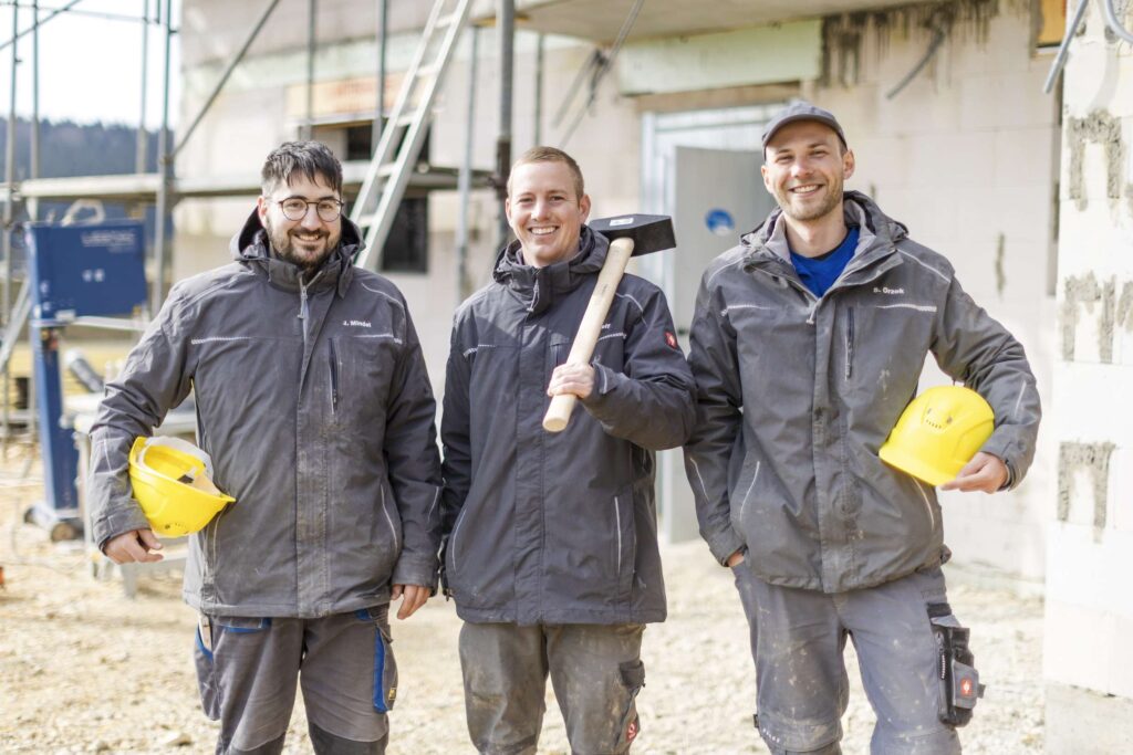 Drei Bauarbeiter von Rudolf Bau Obermarchtal laecheln vor einer Baustelle in die Kamera.