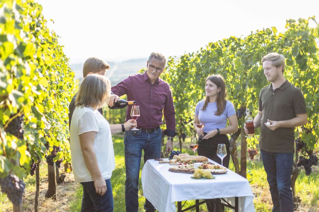 Eine Gruppe von Menschen bei einer Weinprobe im Weinberg, mit einem gedeckten Tisch im Sonnenschein.