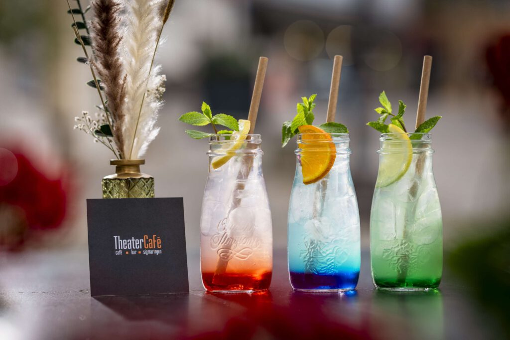 Bunte Cocktails in Flaschen mit floralem Gesteck im Hintergrund.