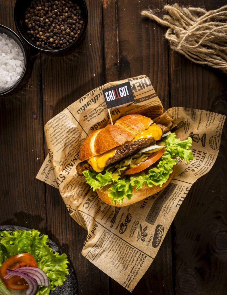 Alt-Text: Ein Burger mit Salat, Tomaten und Zwiebeln auf einem dunklen Holztisch serviert, eingewickelt in Zeitungspapier.