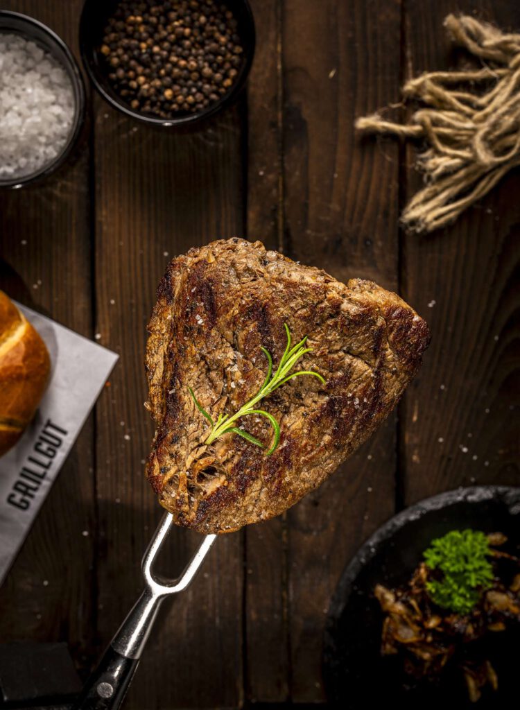 Gegrilltes Steak mit Rosmarin auf einem Holztisch mit Gewuerzen und Brot im Hintergrund.